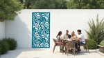 The 'Jungle Mirage' metal privacy screen featuring an intricate swirling leaf pattern, finished in Wedgwood Satin powder coating. The screen is mounted on a white wall in an outdoor patio area, where a group of five people are gathered around a dining table enjoying a meal together, adding elegance and subtle privacy to the space.