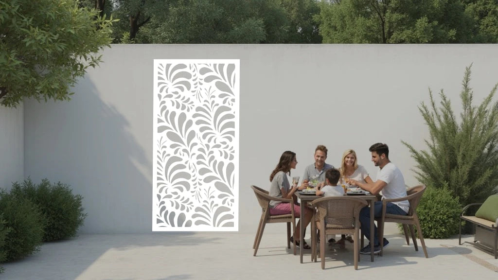 The 'Jungle Mirage' metal privacy screen featuring an intricate swirling leaf pattern, finished in Bright White Gloss powder coating. The screen is mounted on a white wall in an outdoor patio area, where a group of five people are gathered around a dining table enjoying a meal together, adding elegance and subtle privacy to the space