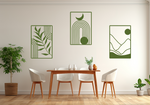 A three-piece minimalist geometric metal wall art set featuring abstract shapes, including a leaf design, a rainbow with moon and sun, and a mountain landscape, all finished in Mistletoe powder coating. The artwork is mounted on a dining room wall above a simple wooden table with white chairs, bringing a modern and artistic touch to the space.