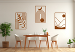 A three-piece minimalist geometric metal wall art set featuring abstract shapes, including a leaf design, a rainbow with moon and sun, and a mountain landscape, all finished in Rust Patina powder coating. The artwork is mounted on a dining room wall above a simple wooden table with white chairs, bringing a modern and artistic touch to the space.