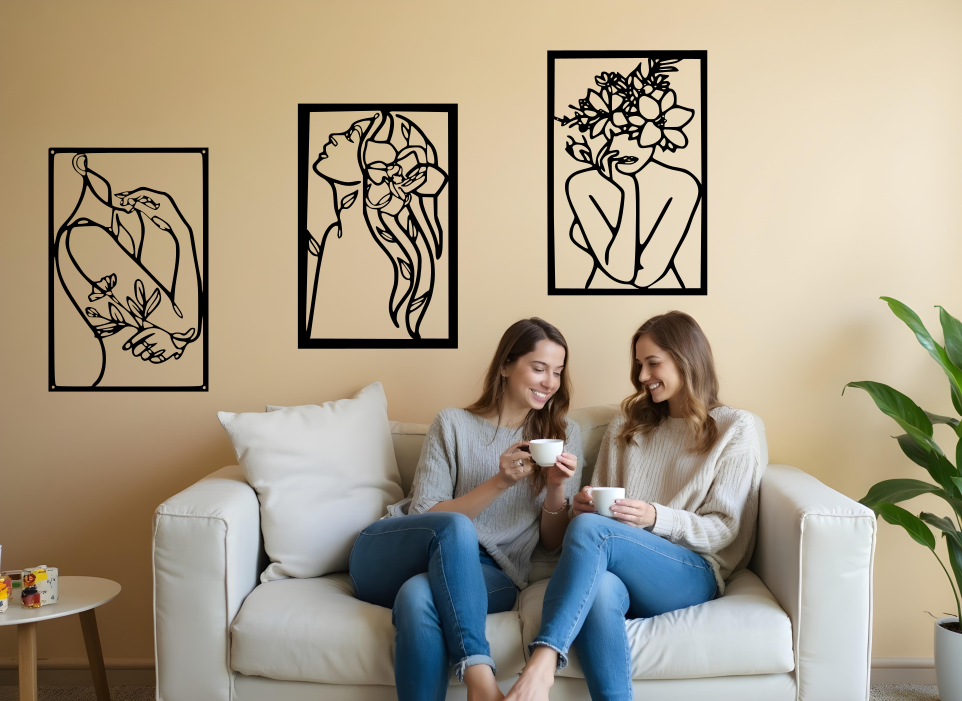 Set of three minimalist metal wall art pieces depicting floral-inspired female figures in various poses, finished in Night Sky Matt Black powder coating, mounted on a neutral wall in a cozy living room setting where two women are enjoying coffee together