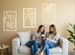 Set of three minimalist metal wall art pieces depicting floral-inspired female figures in various poses, finished in Canola Cream powder coating, mounted on a neutral wall in a cozy living room setting where two women are enjoying coffee together.