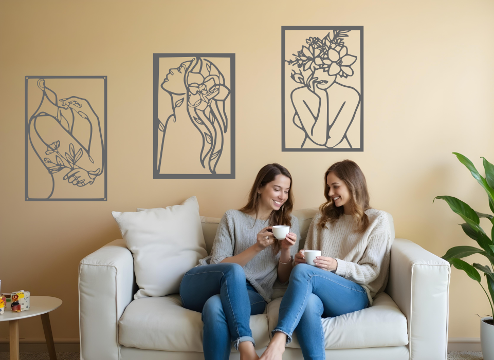 Set of three minimalist metal wall art pieces depicting floral-inspired female figures in various poses, finished in Berry Grey Gloss powder coating, mounted on a neutral wall in a cozy living room setting where two women are enjoying coffee together.