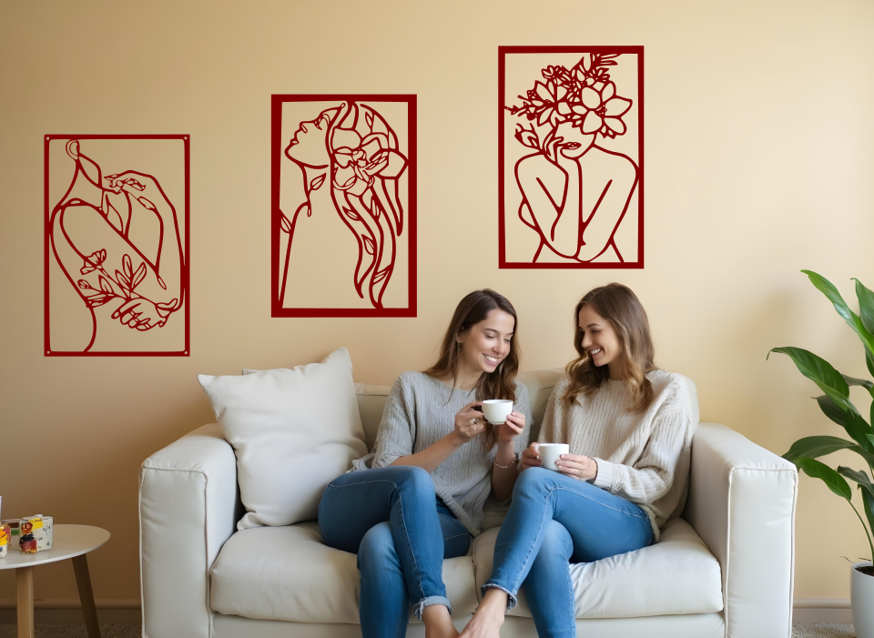 Set of three minimalist metal wall art pieces depicting floral-inspired female figures in various poses, finished in Manor Red powder coating, mounted on a neutral wall in a cozy living room setting where two women are enjoying coffee together.