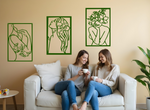 Set of three minimalist metal wall art pieces depicting floral-inspired female figures in various poses, finished in Mistletoe powder coating, mounted on a neutral wall in a cozy living room setting where two women are enjoying coffee together.