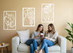 Set of three minimalist metal wall art pieces depicting floral-inspired female figures in various poses, finished in Bright White Gloss powder coating, mounted on a neutral wall in a cozy living room setting where two women are enjoying coffee together.