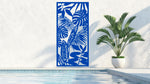 The 'Rainforest Leaves' metal privacy screen featuring an intricate design of tropical leaves, finished in Blaze Blue Gloss powder coating. The screen is mounted on a white wall near a serene poolside area with a large potted plant, adding a vibrant and stylish touch to the outdoor space.